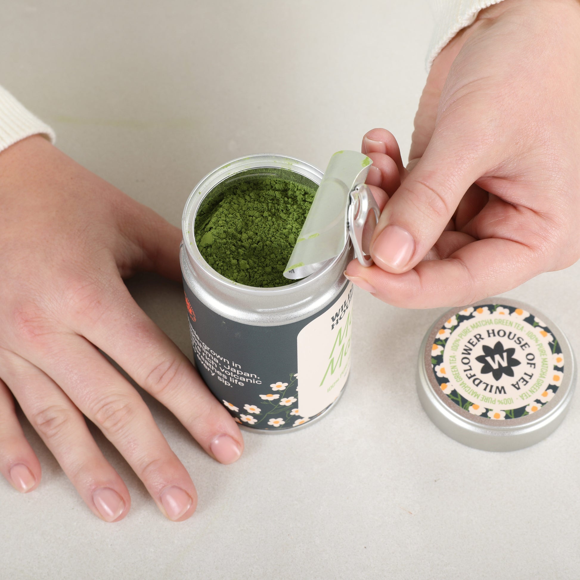 Person opening a container of green powder with a spoon, branded 'Flower House of Wisteria'.